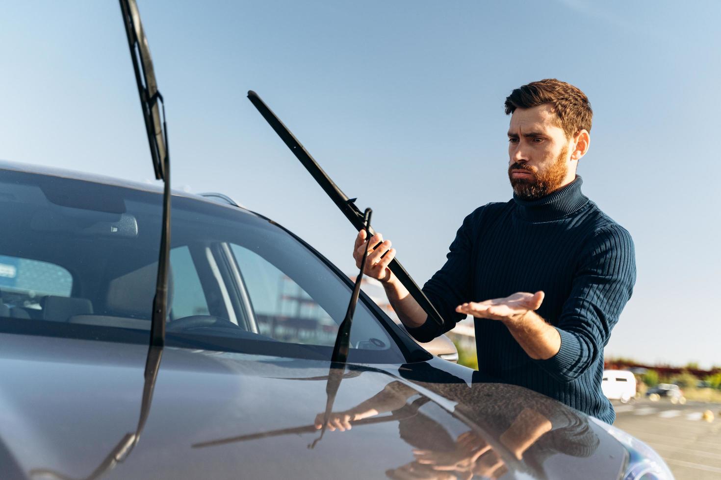 Caucasian man feeling confused while changing windscreen wipers on a car at the street during the sunny day. Transportation concept photo