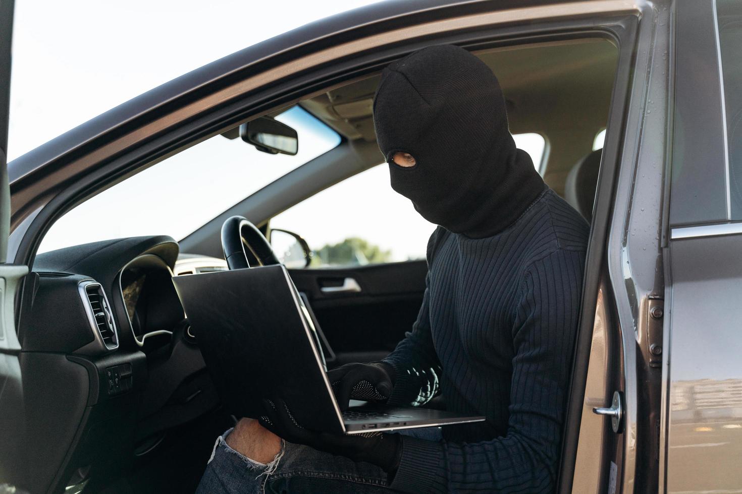 ladrón enmascarado con un pasamontañas robando un portátil del coche y mirando la pantalla mientras está sentado dentro. concepto criminal. foto de stock