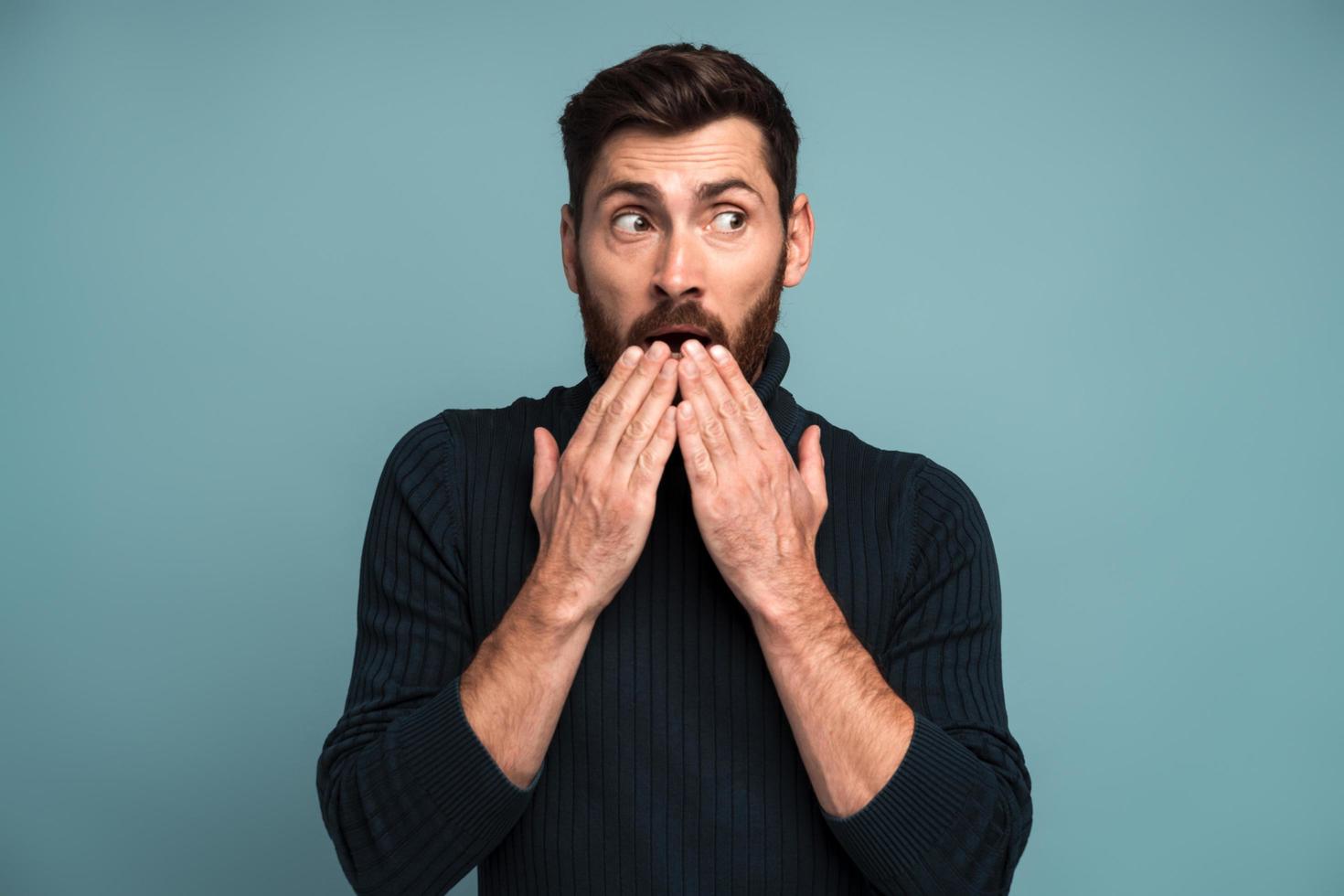 Wow. Man covering mouth with hand and looking away with big eyes, scared surprised expression, shocked by sudden news. Indoor studio shot isolated on blue background photo