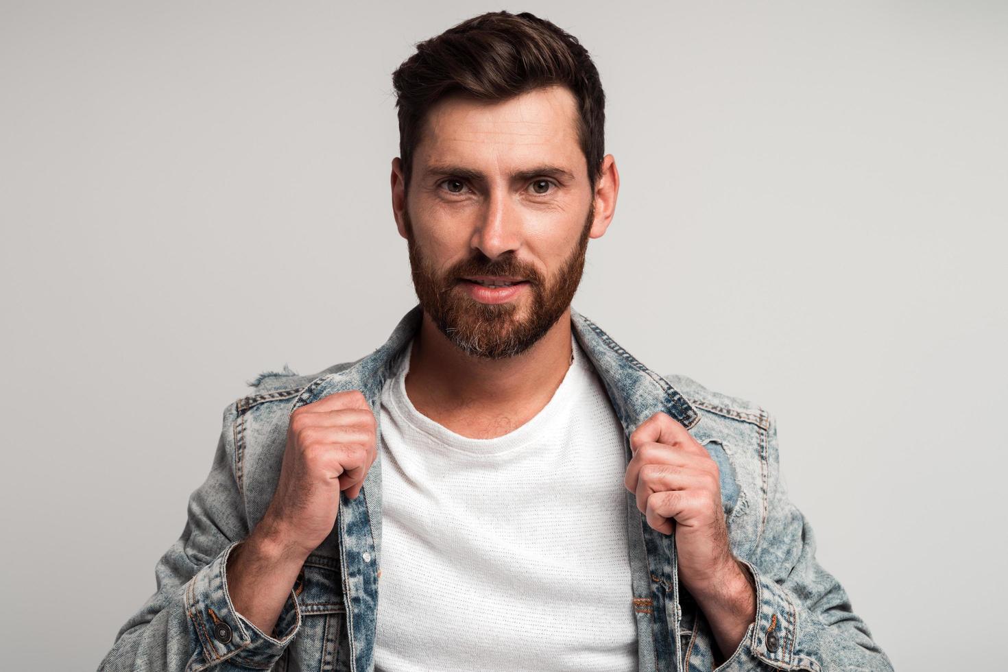 Retrato de hombre guapo con barba en ropa casual mirando a la cámara y sonriendo mientras corrige su chaqueta y posa. Foto de estudio con fondo blanco