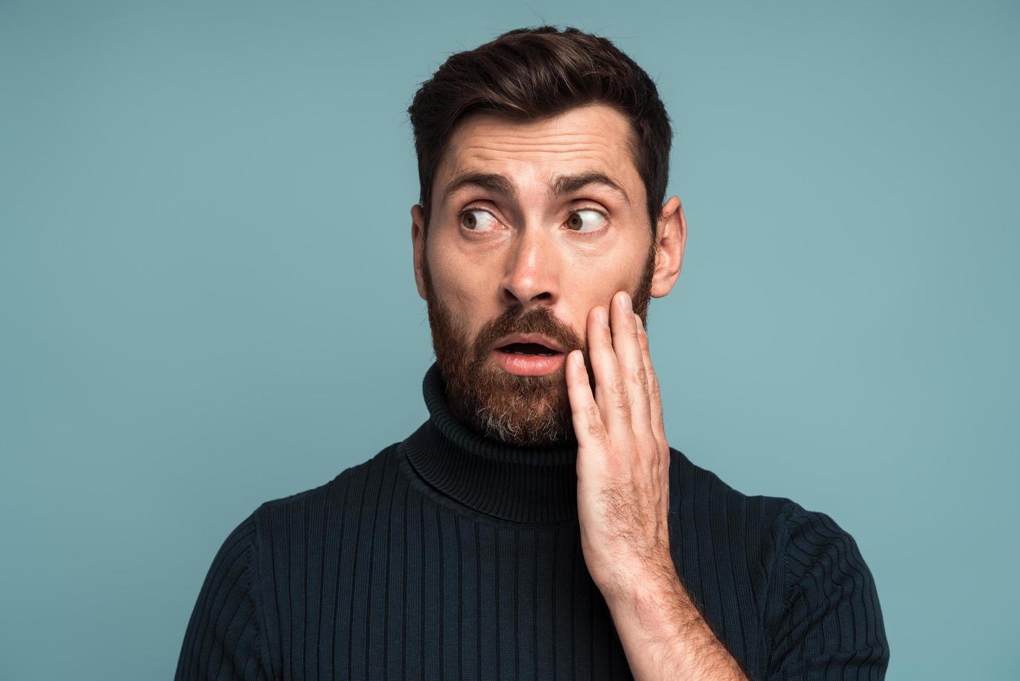 Excited shocked man holding hands on face and looking away with big eyes and open mouth, shocked bemused with news, win at lottery. Indoor studio shot isolated on blue background photo