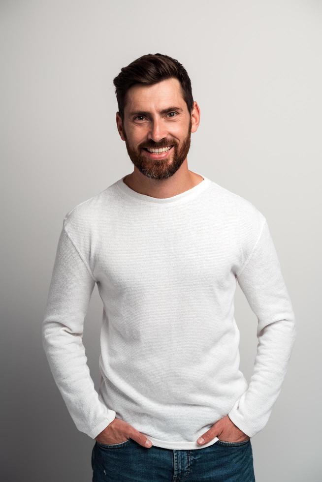 Portrait of happy satisfied handsome young man in white shirt standing with arms at the pockets and looking at camera with toothy smile. Indoor studio shot on white background copy space photo