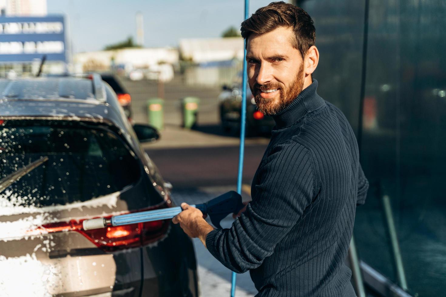 Waist up portrait view of the caucasian man holding special equipment and smiling to the camera while cleaning car using high pressure water. Car washing concept. Selective focus photo