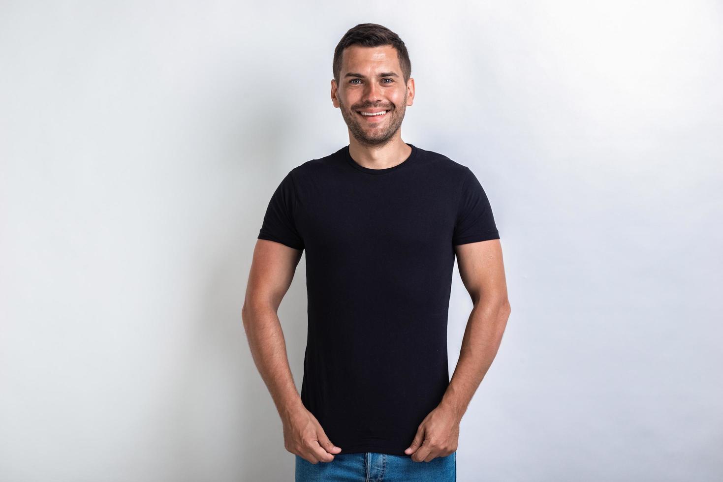 Hombre divertido vestido con camiseta negra de pie tirando hacia abajo una camiseta sonriendo y mirando a la cámara.- imagen foto