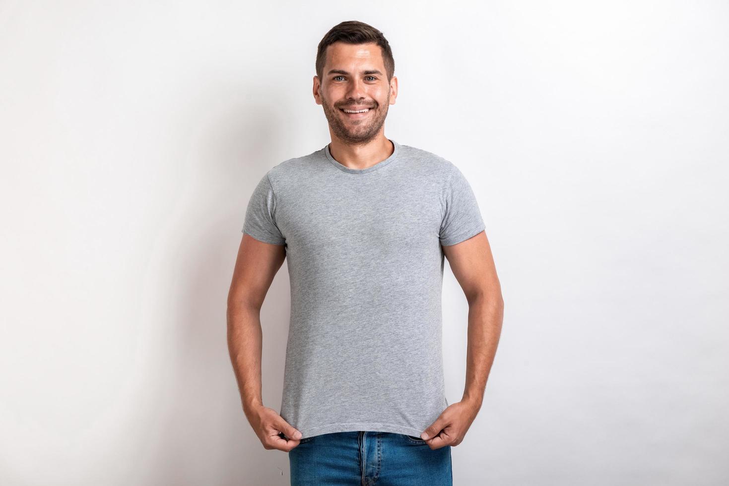 Hombre divertido vestido con camiseta gris de pie tirando hacia abajo una camiseta sonriendo y mirando a la cámara.- imagen foto