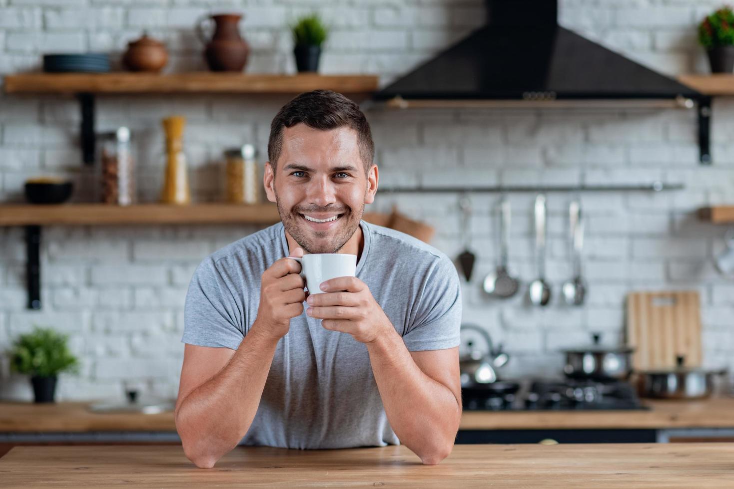 Hombre guapo se sienta en la mesa de la cocina con una taza de té o café por la mañana y sonriendo mirando a la cámara foto