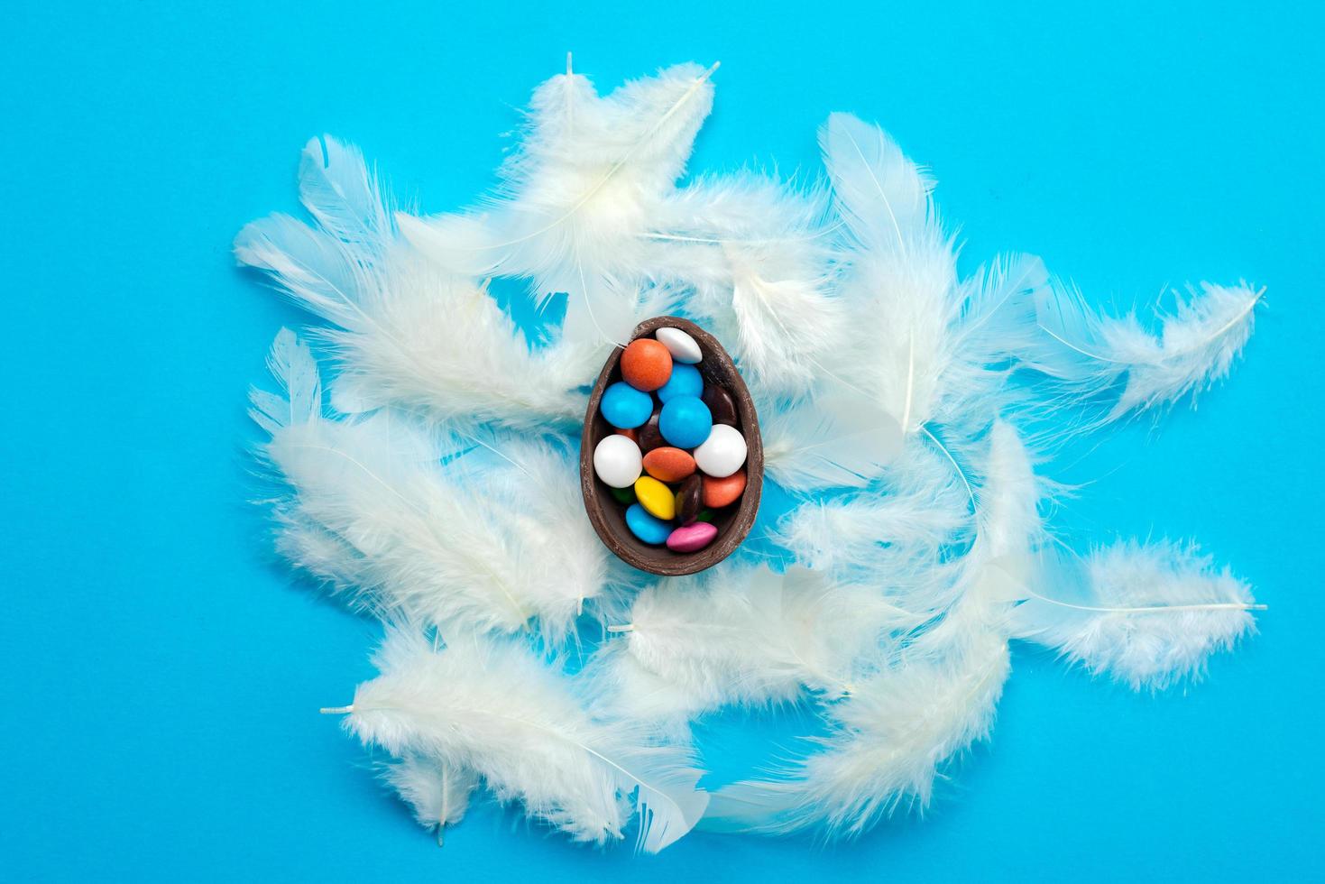 White feathers  lie on the blue Easter background. Multi-colored candies in the chocolate egg photo