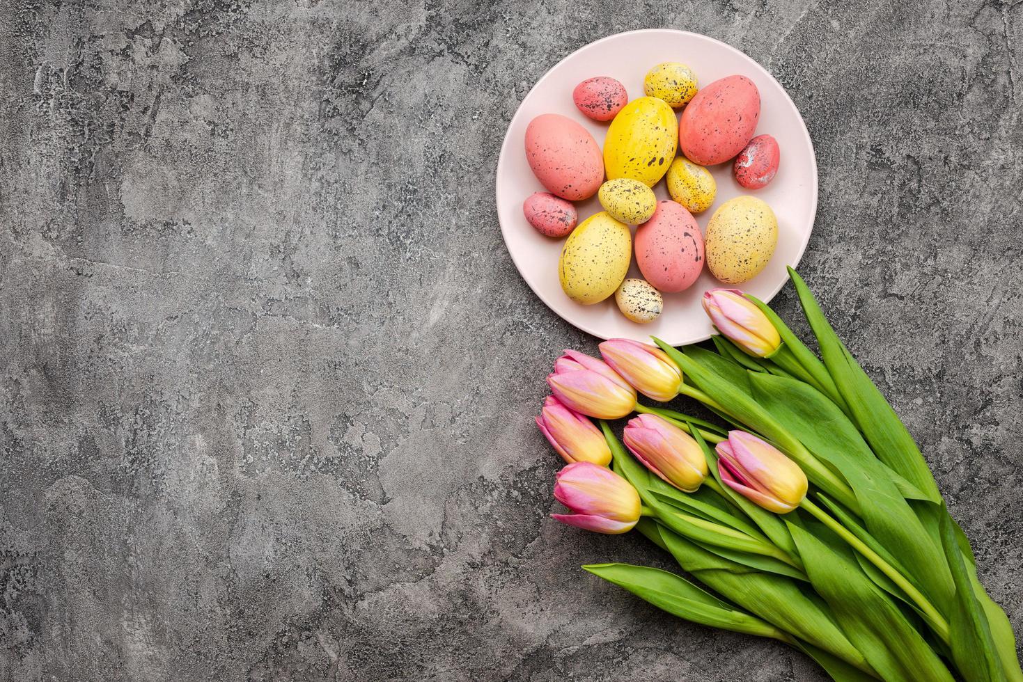 sobre un fondo de yeso gris hay hermosos tulipanes y un plato con coloridos huevos de pascua. copia espacio foto