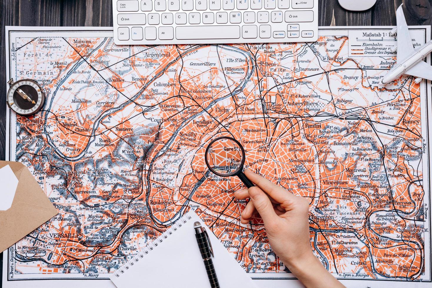 A map is laid out on a wooden table, a man is looking at something from a magnifying glass, a compass, a notebook and a pen photo