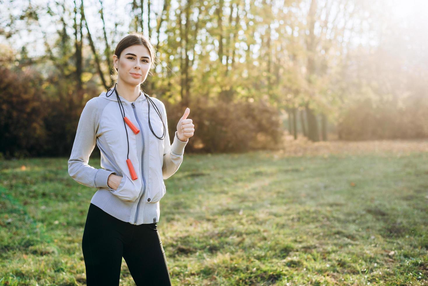 Beautiful girl with skipping rope on shoulder outdoors showing thumb up. photo