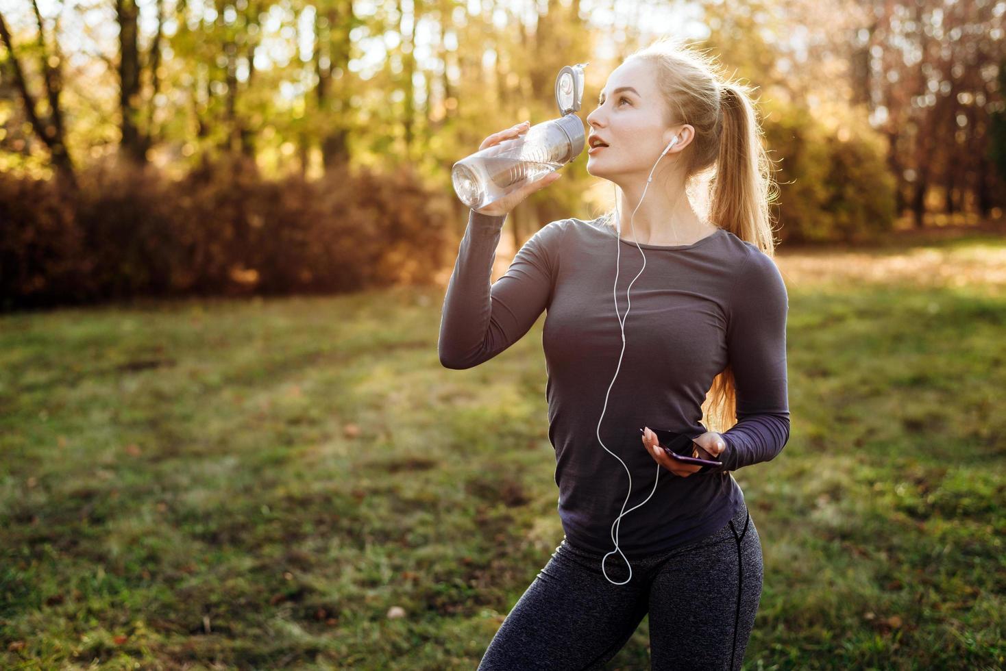 Fitness in the park, girl drinking water, holding smartphone and headphones in her hand. photo