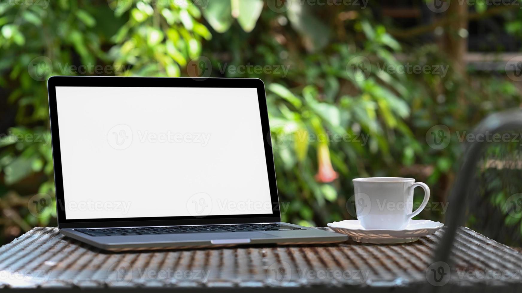 maqueta de pantalla en blanco portátil y taza de café con leche en la mesa de hierro en el jardín, fondo de árbol verde. foto