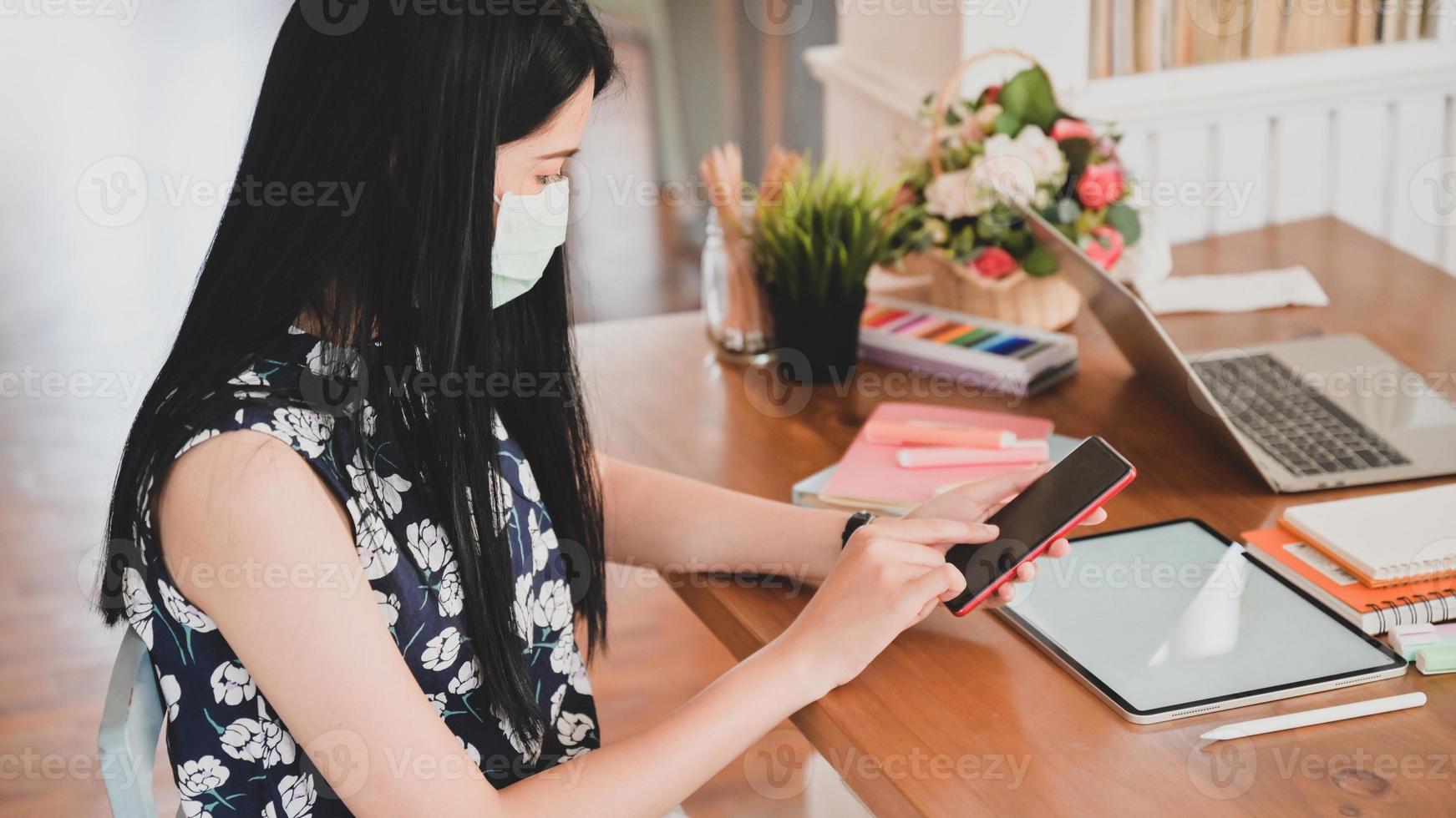 mujer que usa una máscara médica está usando un teléfono inteligente con una tableta, computadora portátil y equipo de oficina en el escritorio. foto