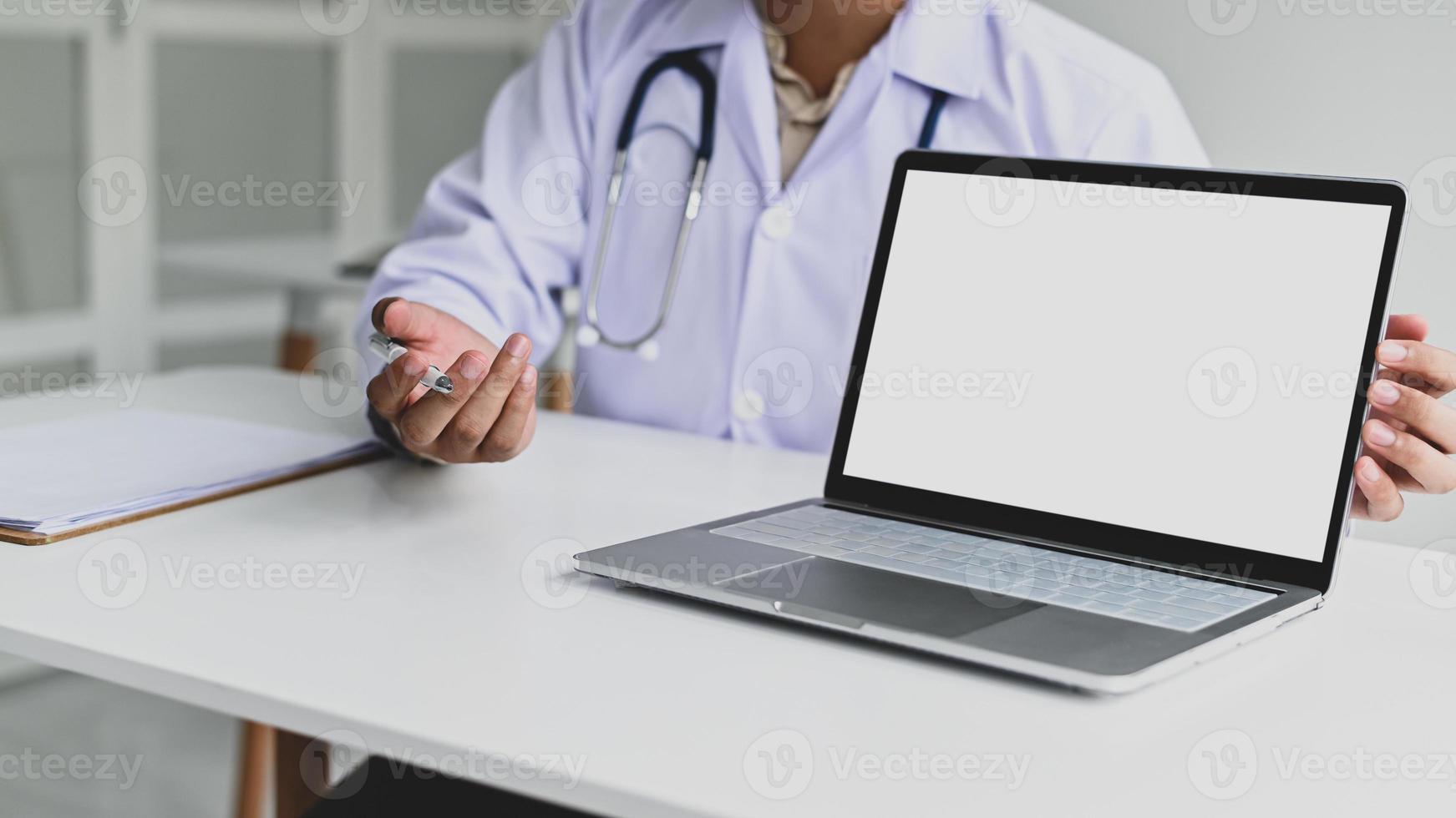 un hombre vestido con una bata médica, sostiene un bolígrafo con una pantalla de computadora portátil en blanco al frente. foto