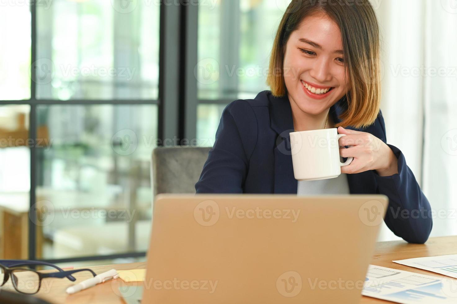 una mujer joven con un traje con una taza de café en la mano está sonriendo y mirando su computadora portátil. foto