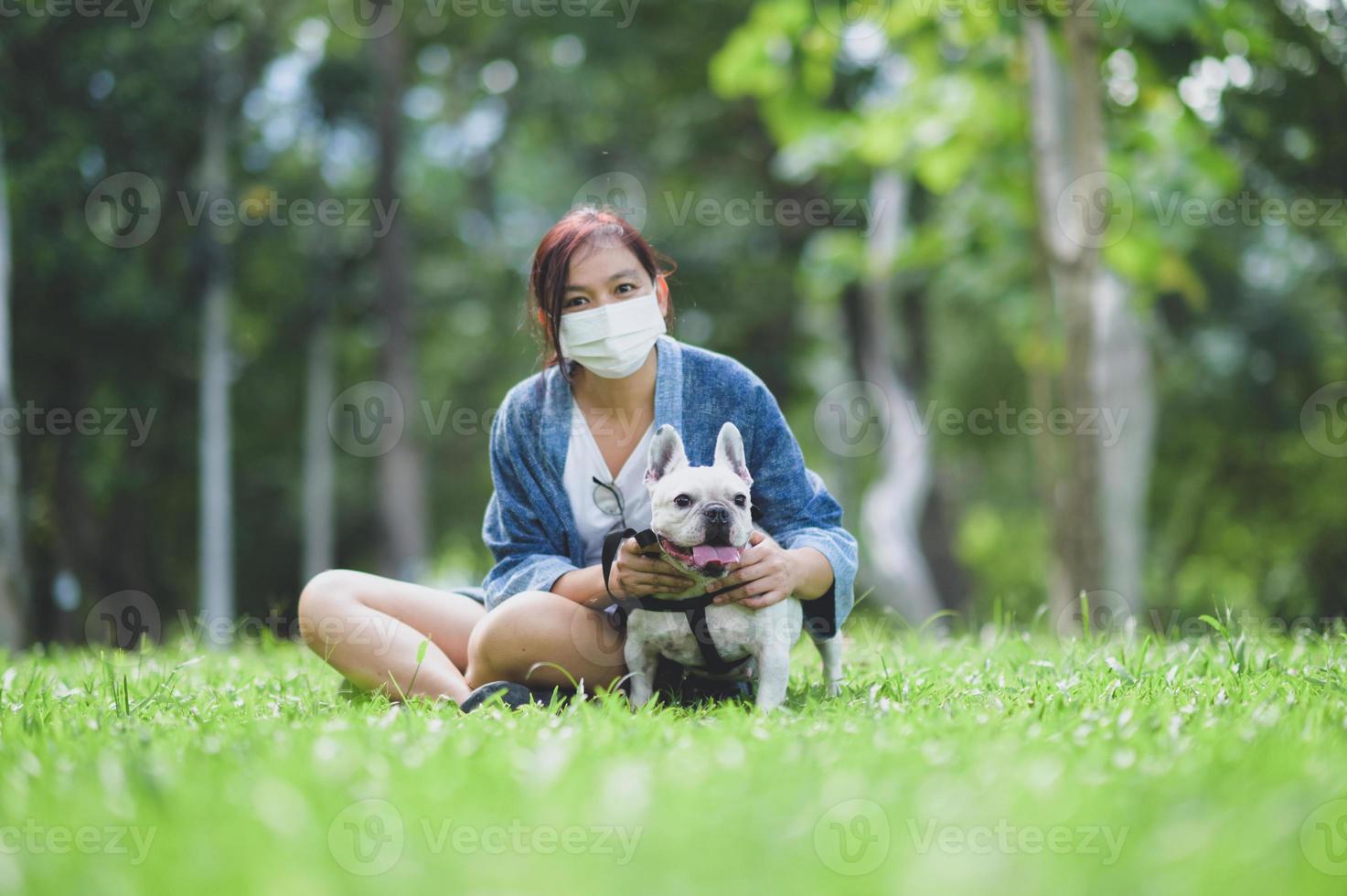 A woman wearing a medical mask sitting on the lawn with a white French bulldog. photo