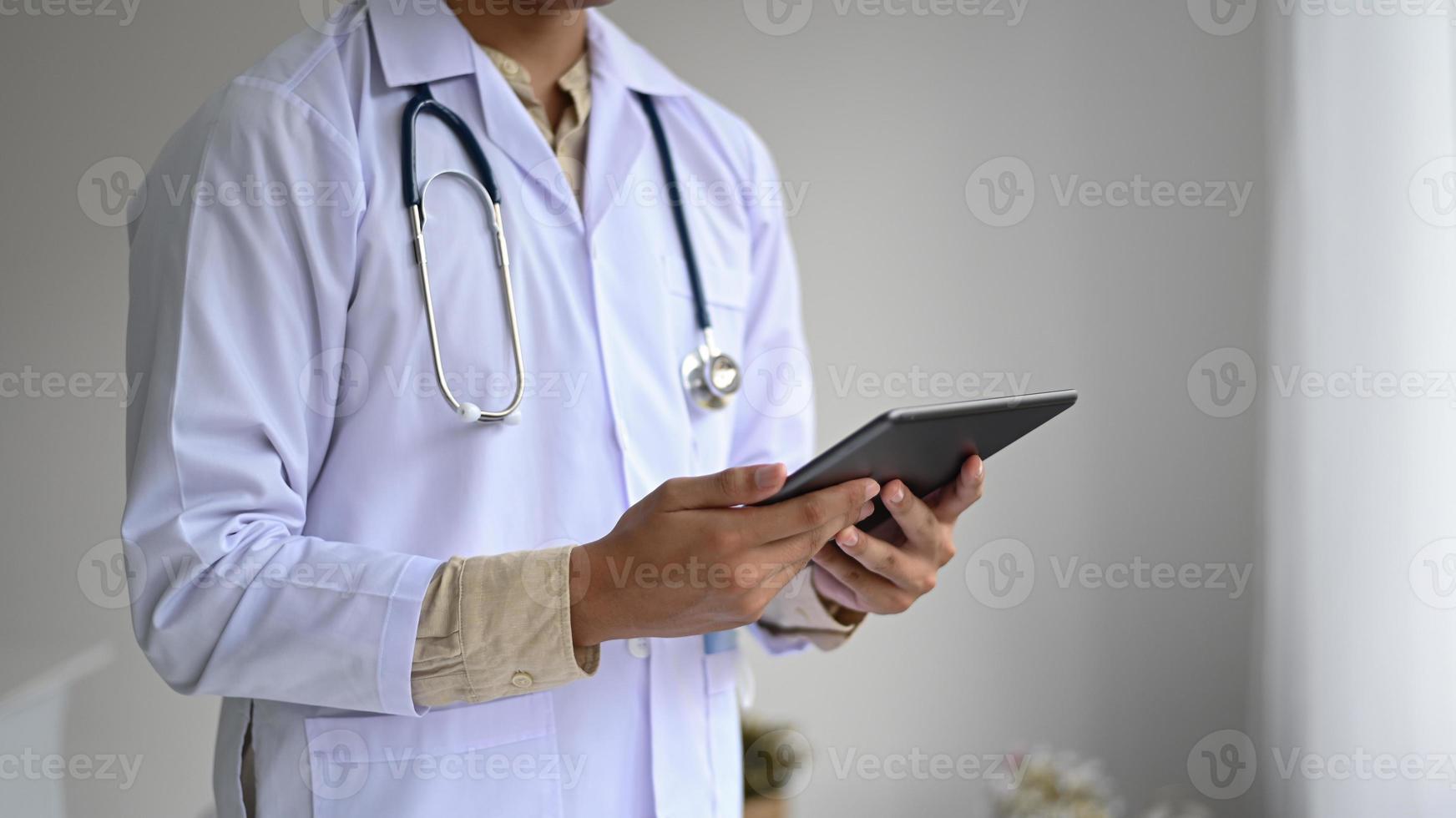 Toma recortada de un profesional médico en una bata de laboratorio con un estetoscopio sosteniendo una tableta en la mano, tiro de vista lateral. foto