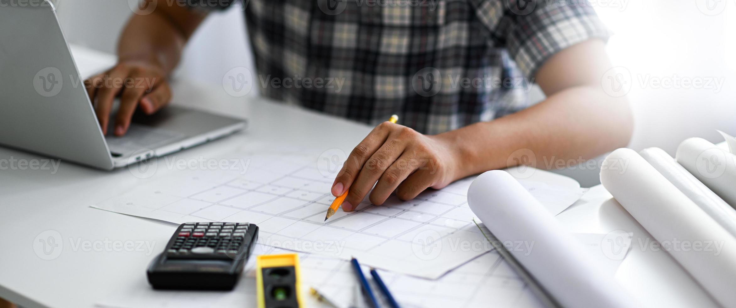 The architect is pointing a pencil at a house plan and working on a laptop. He is checking and recording the results. photo