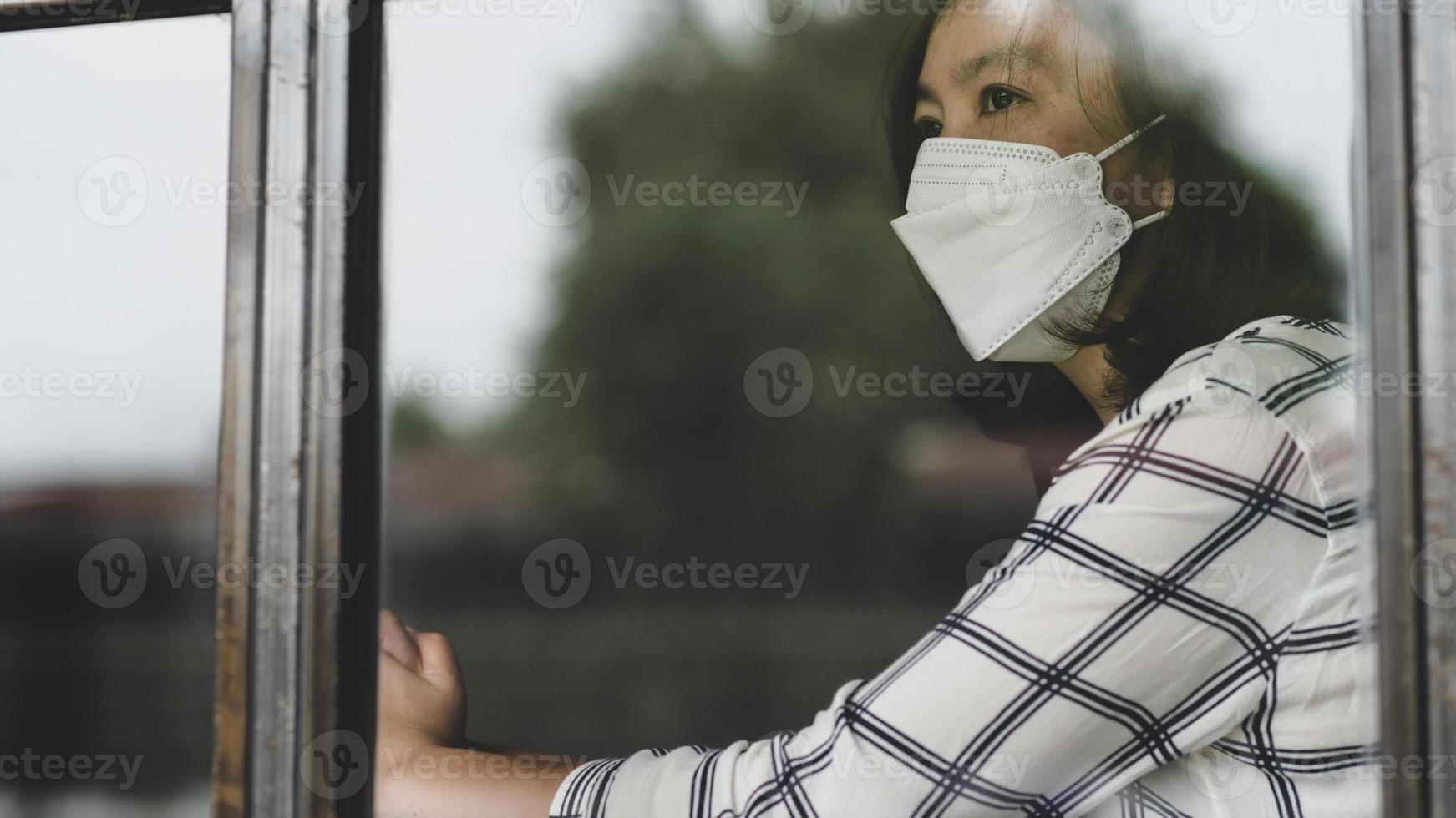 mujer asiática con una máscara médica mirando por la ventana de su casa, trabajando desde casa, cuarentena. foto