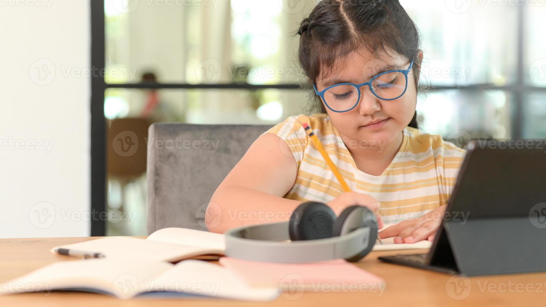 niña asiática con gafas está estudiando en línea, toma notas y se coloca una tableta sobre la mesa. foto