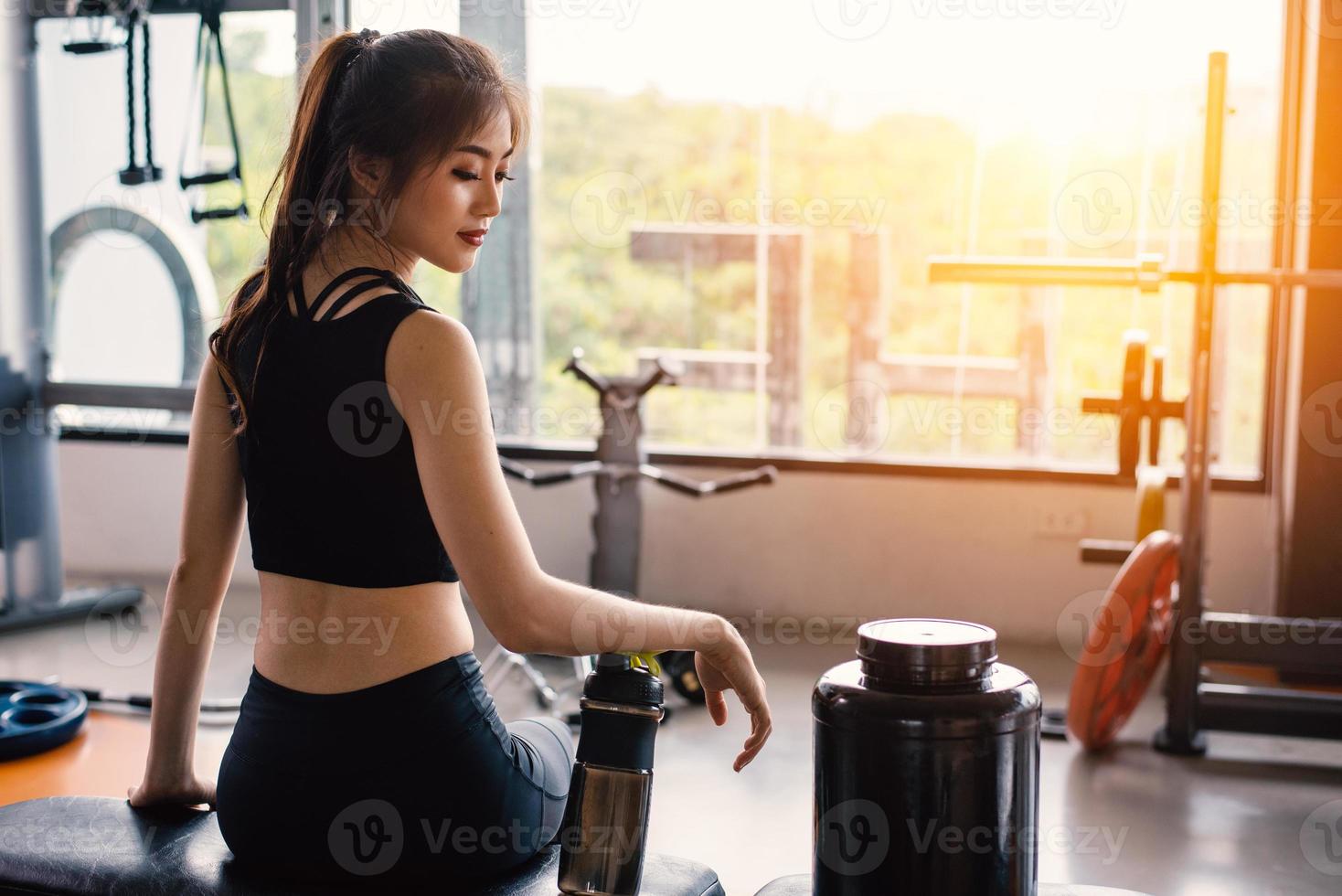 deporte mujer sentada y descansando después de entrenar o hacer ejercicio en el gimnasio con batido de proteínas o agua potable en el suelo. relajarse concepto. tema de entrenamiento de fuerza y desarrollo corporal. tono cálido y fresco foto