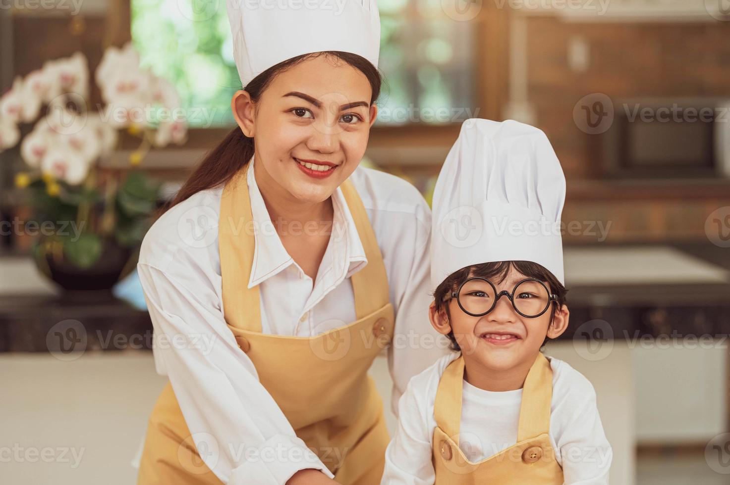 Portrait cute little Asian boy chef with eyeglasses and his mother looking to camera in home cooking kitchen happily and funny. People lifestyles and Family. Homemade food ingredient making concept photo