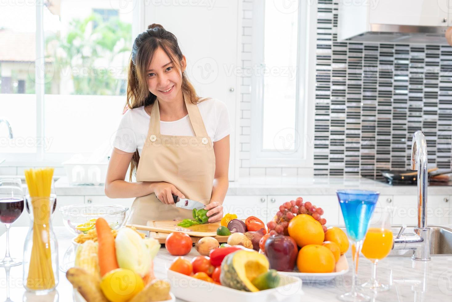 Asian beauty woman cooking and slicing vegetable in kitchen room with full of food and fruit on table for party. Holiday and Happiness concept. People and lifestyles concept. Girl looking at camera photo