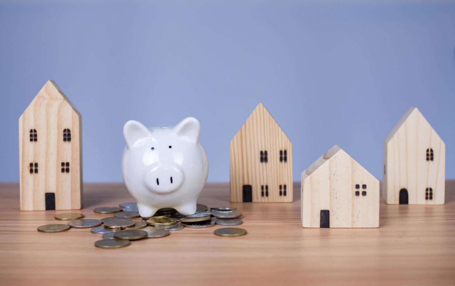 A white piggy bank is placed on a pile of coins. And beside it is a wooden model house on a white background. which are all on a wooden table photo