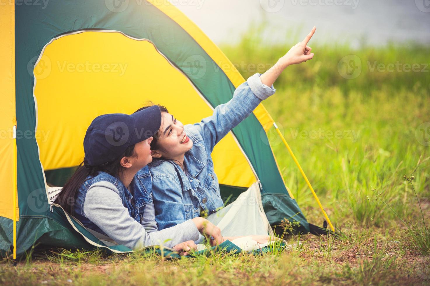 Two close friend Asian friendship relax in camping tent in green meadow on lake side view background. Girl pointing finger to sky. People lifestyle travel on vacation concept. Summer picnic activity photo