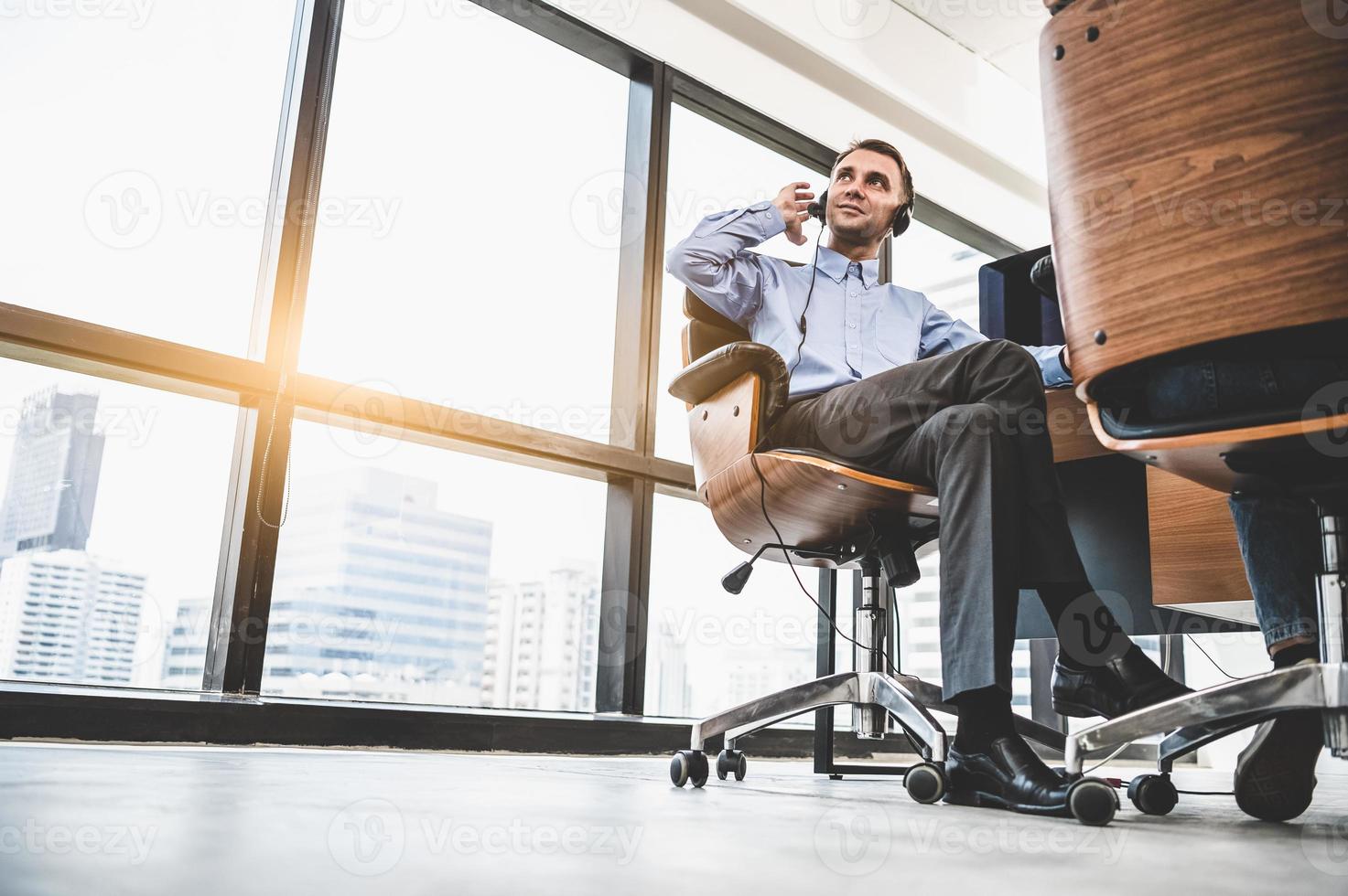 Portrait of happy handsome business man with headset in modern office with city urban building background. Businessman sitting on chair. Caucasian man relaxing in customer care service call center photo