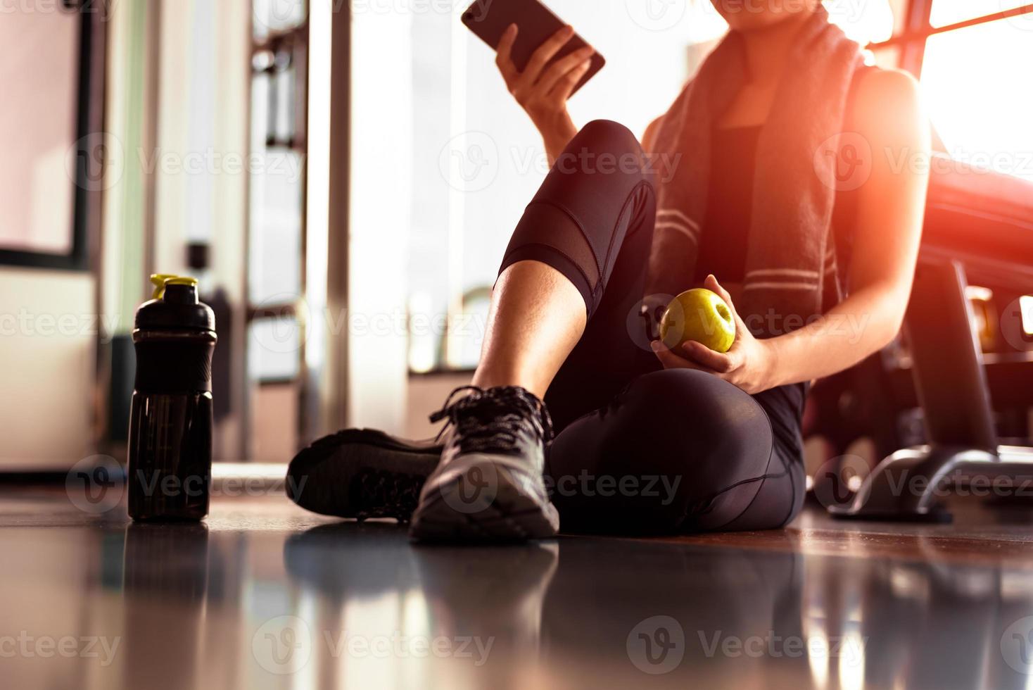 Close up of woman using smart phone and holding apple while workout in fitness gym. Sport and Technology concept. Lifestyles and Healthcare theme photo