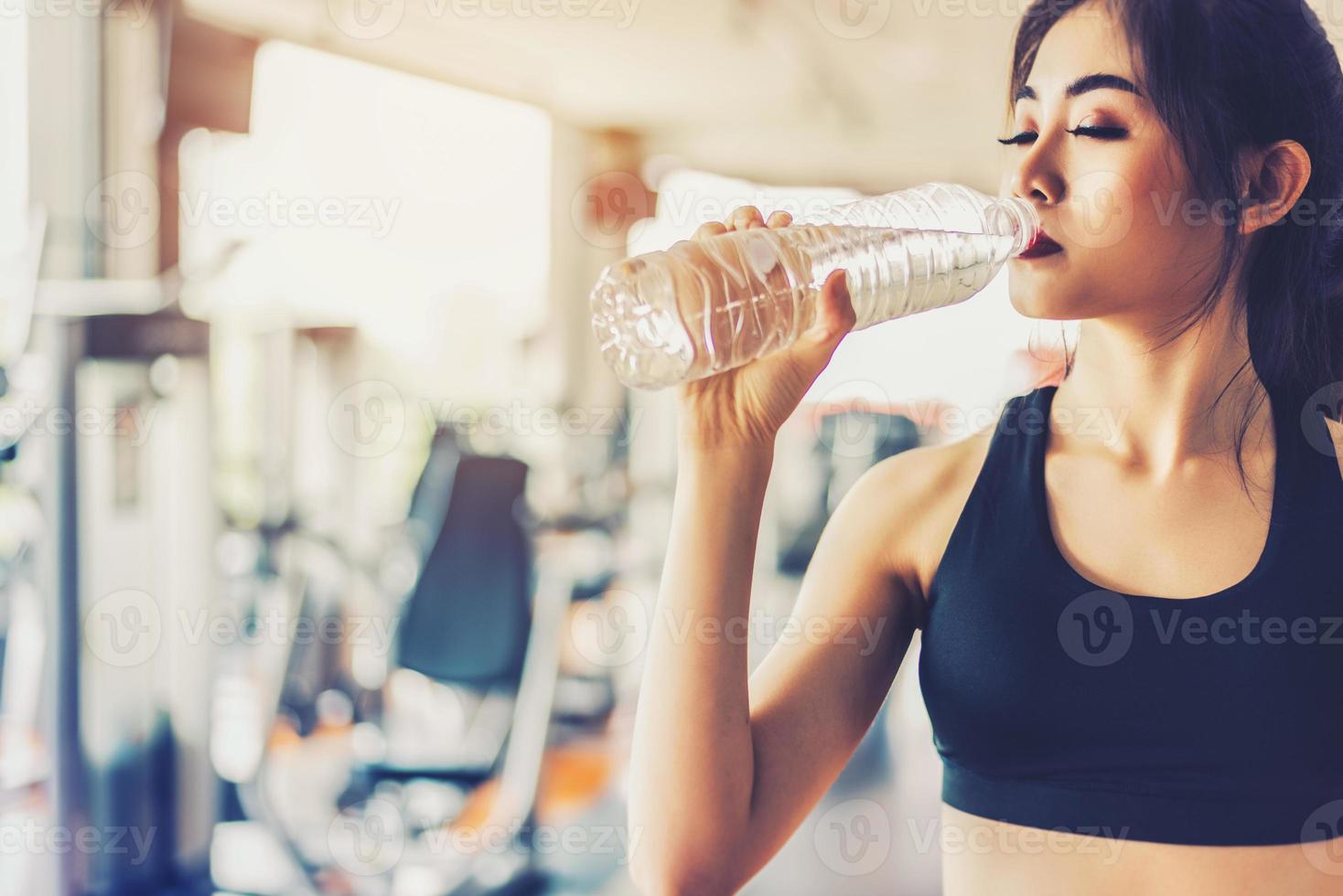 Mujer asiática bebiendo agua potable pura para la frescura después del entrenamiento o el entrenamiento físico en el gimnasio con fondo de equipos de fitness. relajarse y descansar concepto. tema de entrenamiento de fuerza y fitness foto