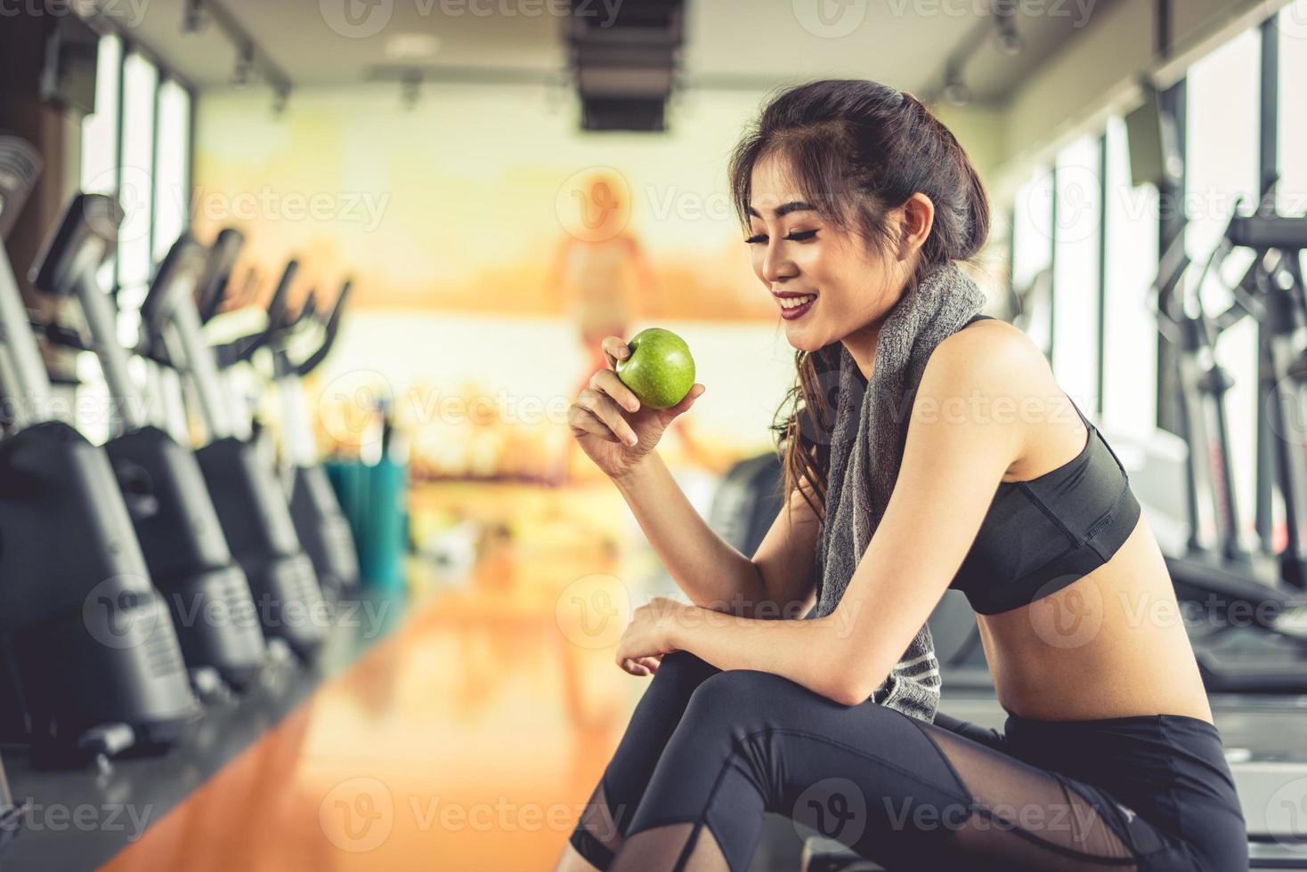 Asian woman holding and looking green apple to eat with sports equipment and treadmill in background. Clean food and Healthy concept. Fitness workout and running theme photo