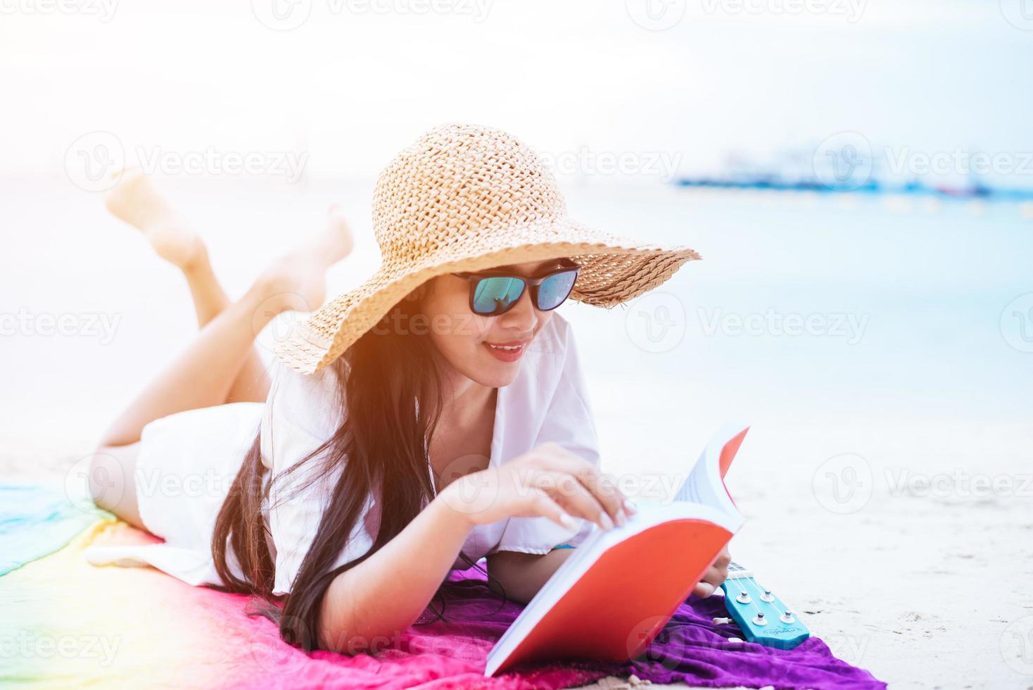 Beauty Asian woman have vacation on beach. Girl wearing wing hat and reading book on colorful mat near sea. Lifestyle and happy life concept. Travel and holiday theme photo