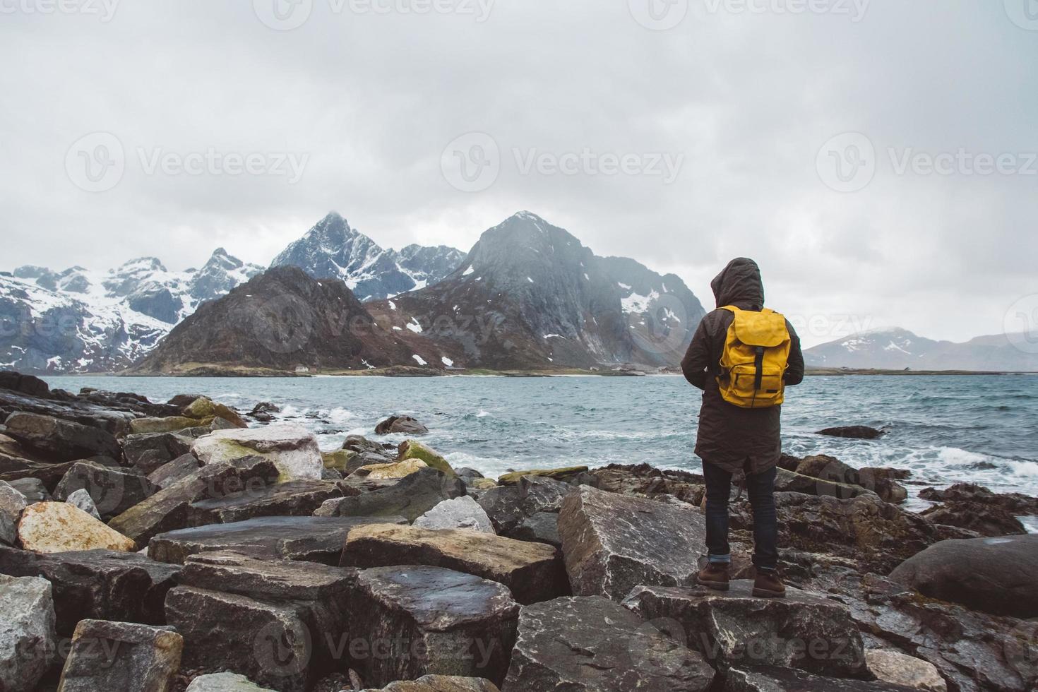 Hombre viajero con una mochila amarilla de pie sobre las rocas contra el fondo del mar y las montañas foto