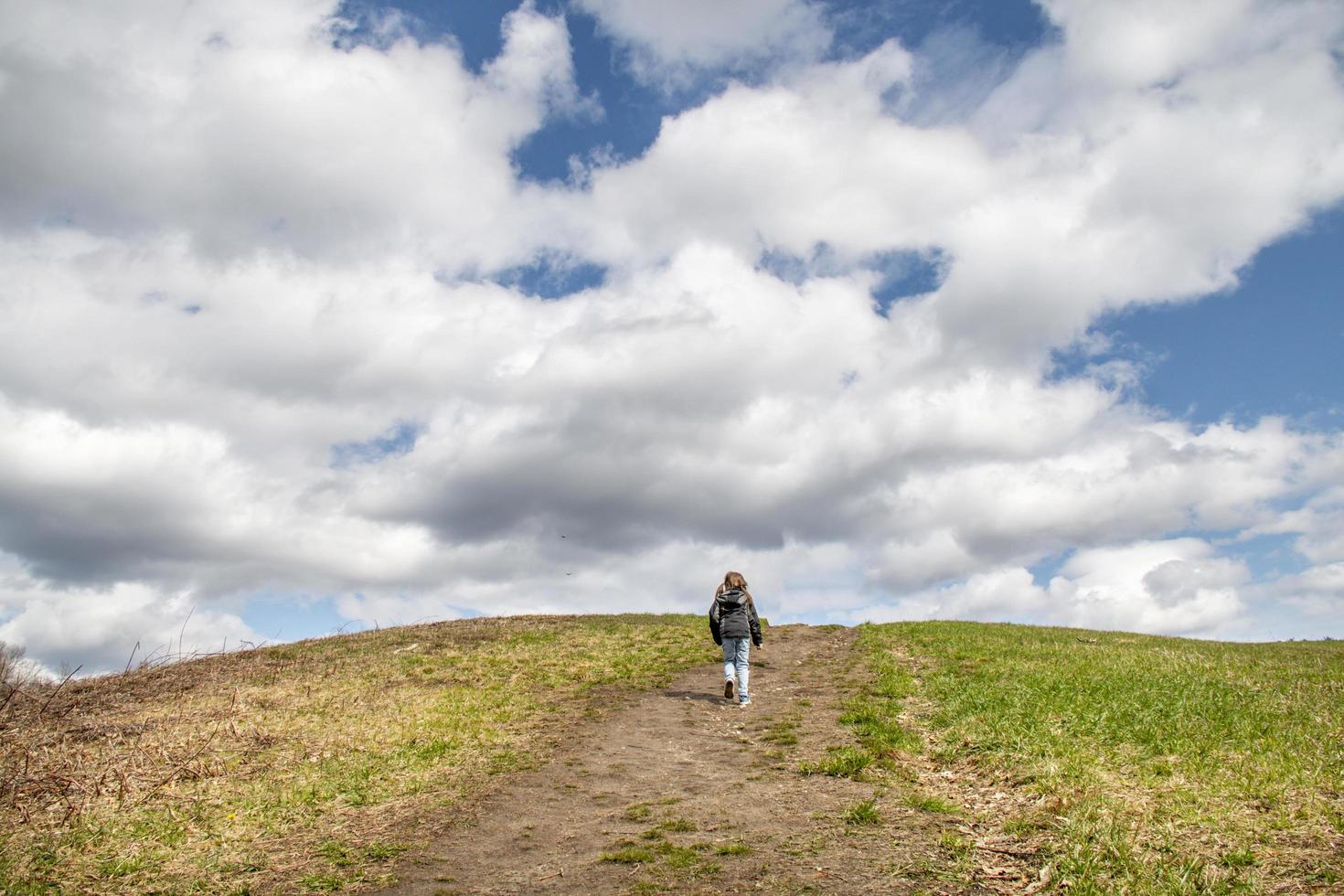 Child walking up a grassy hill toward a cloudy sky photo