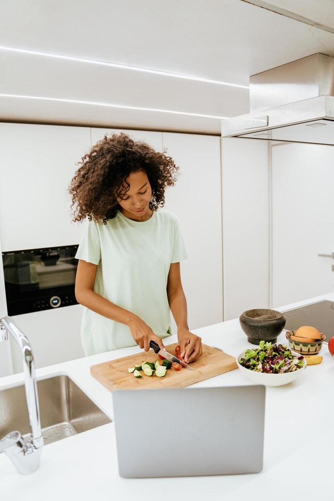 Black young woman making salad at home while using laptop photo
