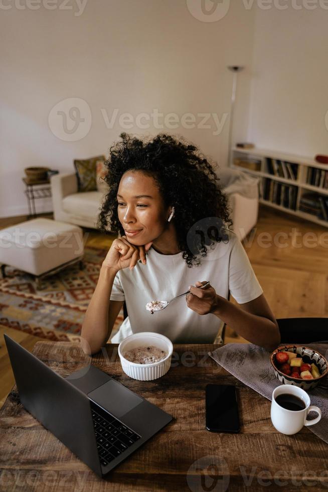 mujer joven negra en auriculares usando la computadora portátil y desayunando foto