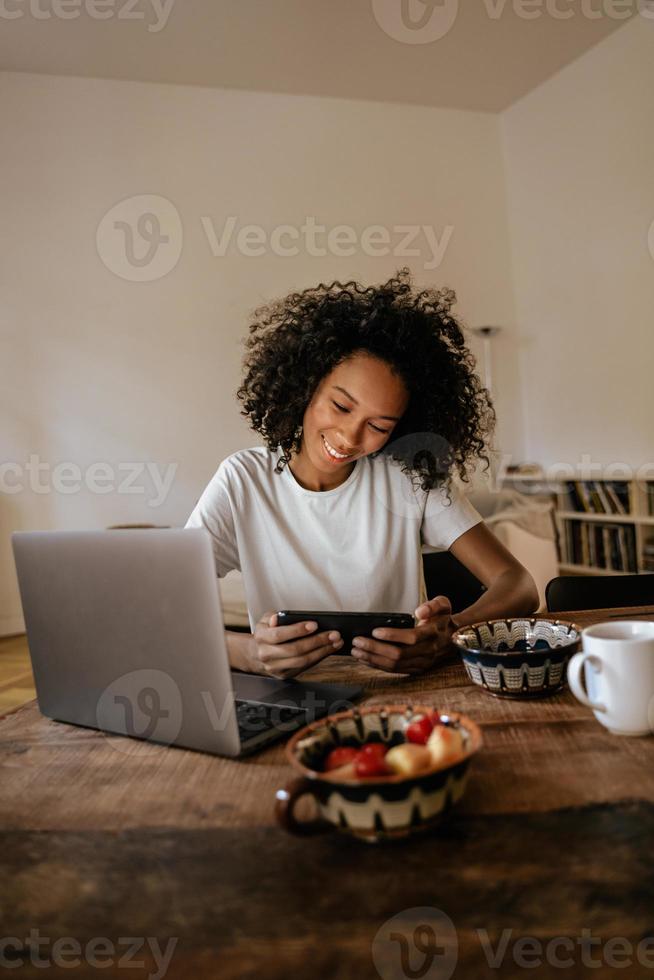 Joven negra con teléfono móvil y portátil mientras desayuna foto