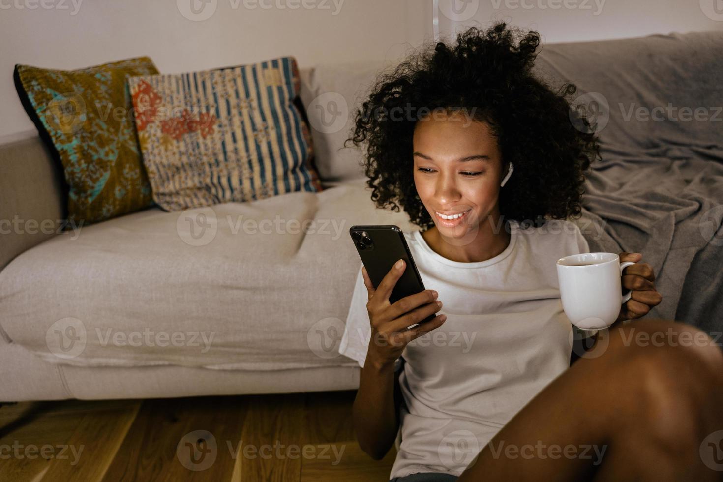 Black young woman in earphones using cellphone and drinking coffee photo