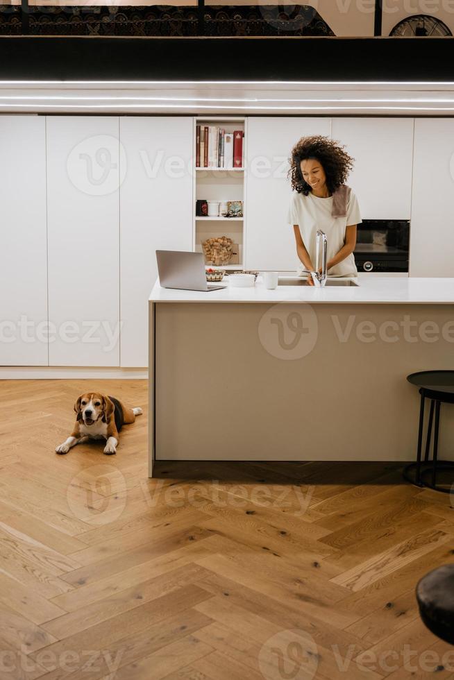 Black young woman washing dishes using her laptop in her kitchen photo