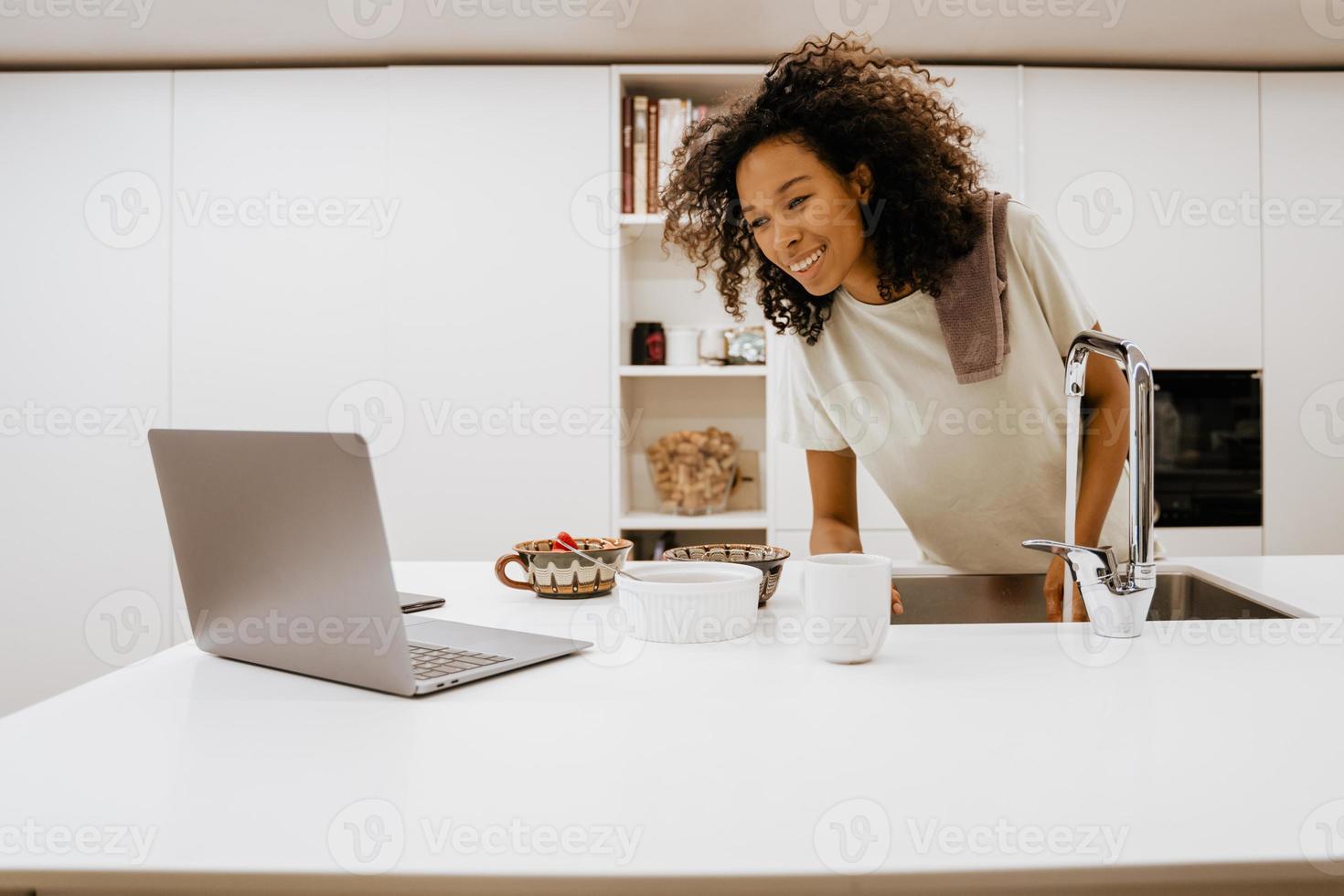 Black young woman washing dishes while using laptop at kitchen photo