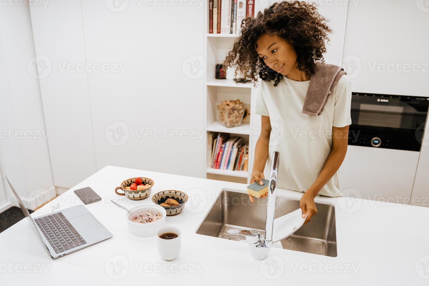 Joven negra lavando platos mientras usa el portátil en la cocina foto