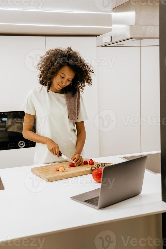 Black young woman making salad while using laptop at kitchen photo