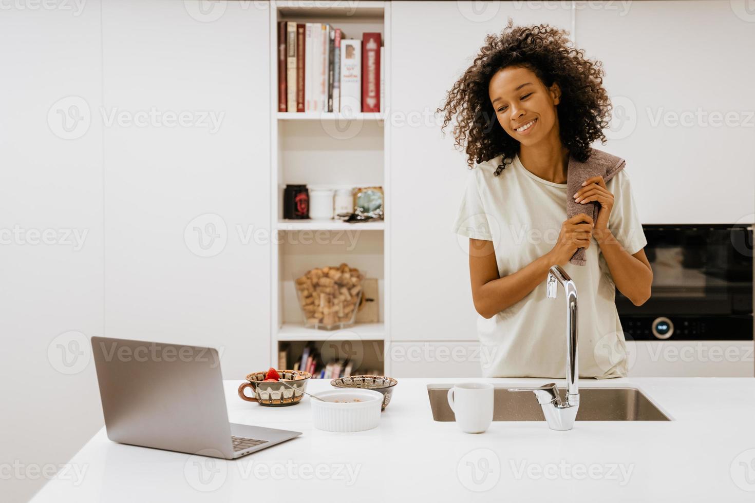 Black young woman washing dishes while looking at her laptop in the kitchen photo