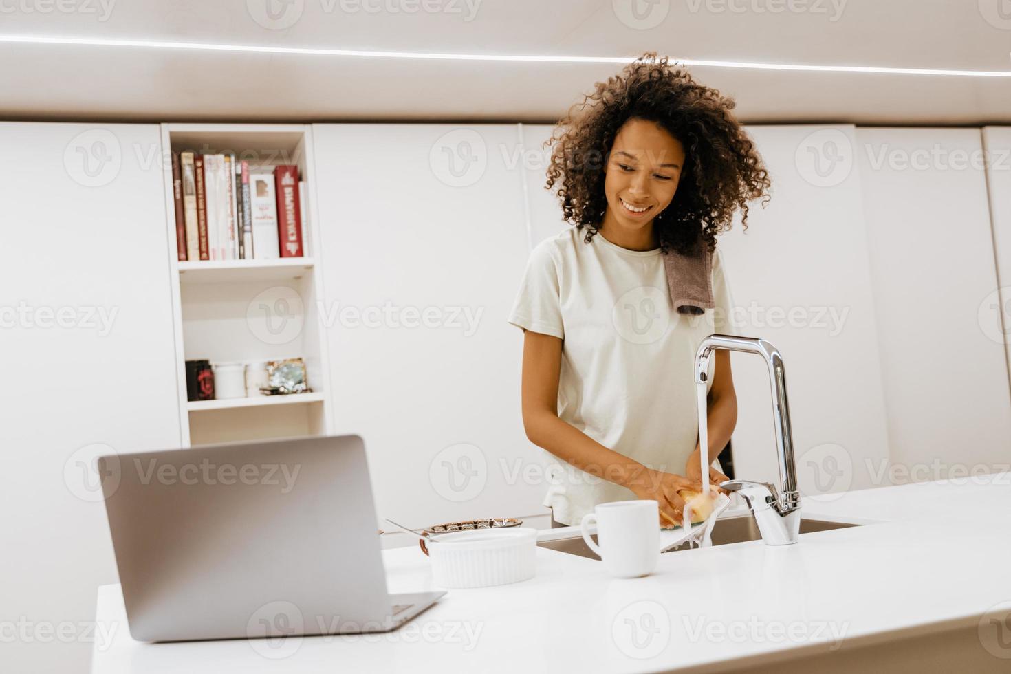 Black young woman washing dishes while using laptop at her kitchen counter photo
