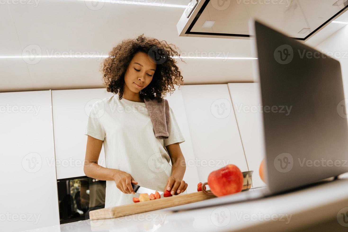 Joven negra haciendo ensalada mientras usa el portátil en la cocina foto