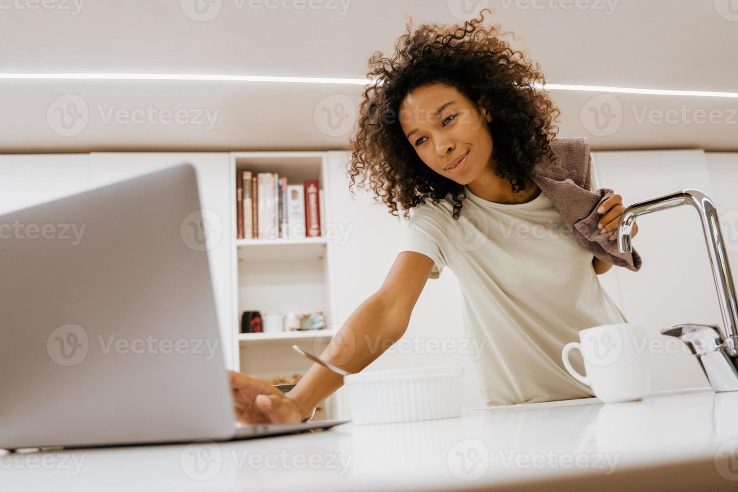 Black young woman washing dishes while using laptop in the kitchen photo