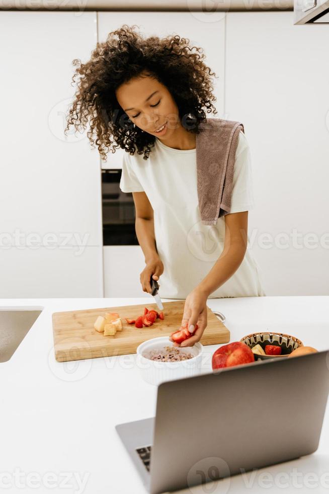 Black young woman making salad while using laptop at kitchen photo