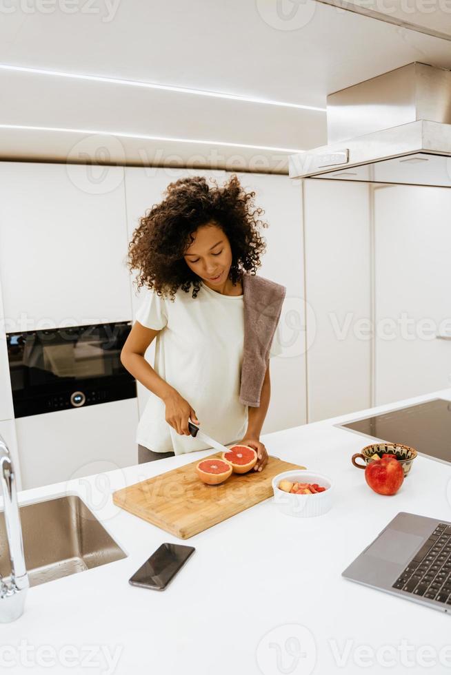Joven negra haciendo el desayuno mientras usa el portátil en la cocina foto